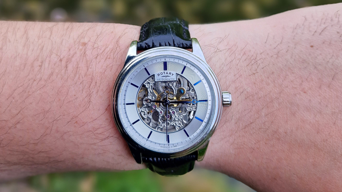 Close-up of a Rotary automatic skeleton watch with a silver case, visible mechanical movement, and black leather strap, worn on a wrist outdoors.