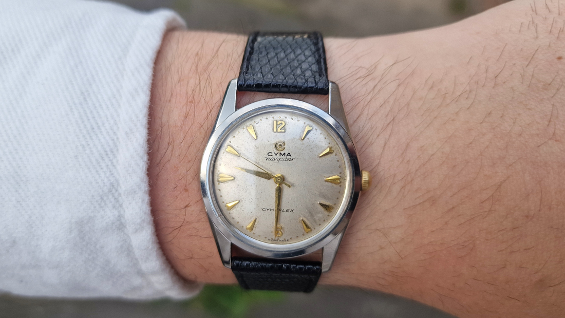 1950s Cyma Navystar wristwatch with champagne dial, gold hour markers, and black leather strap worn on wrist.