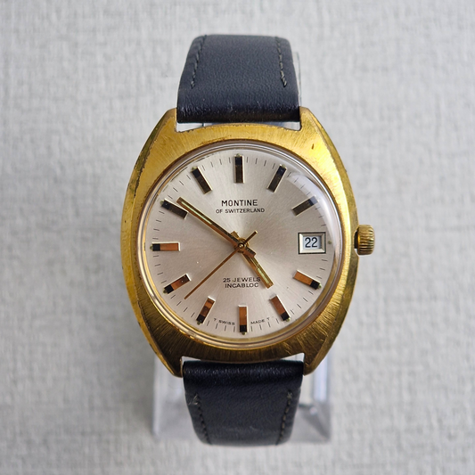 A 1970s Montine of Switzerland gold-plated automatic watch with a champagne dial, black leather strap, and date window, displayed upright on a clear stand.
