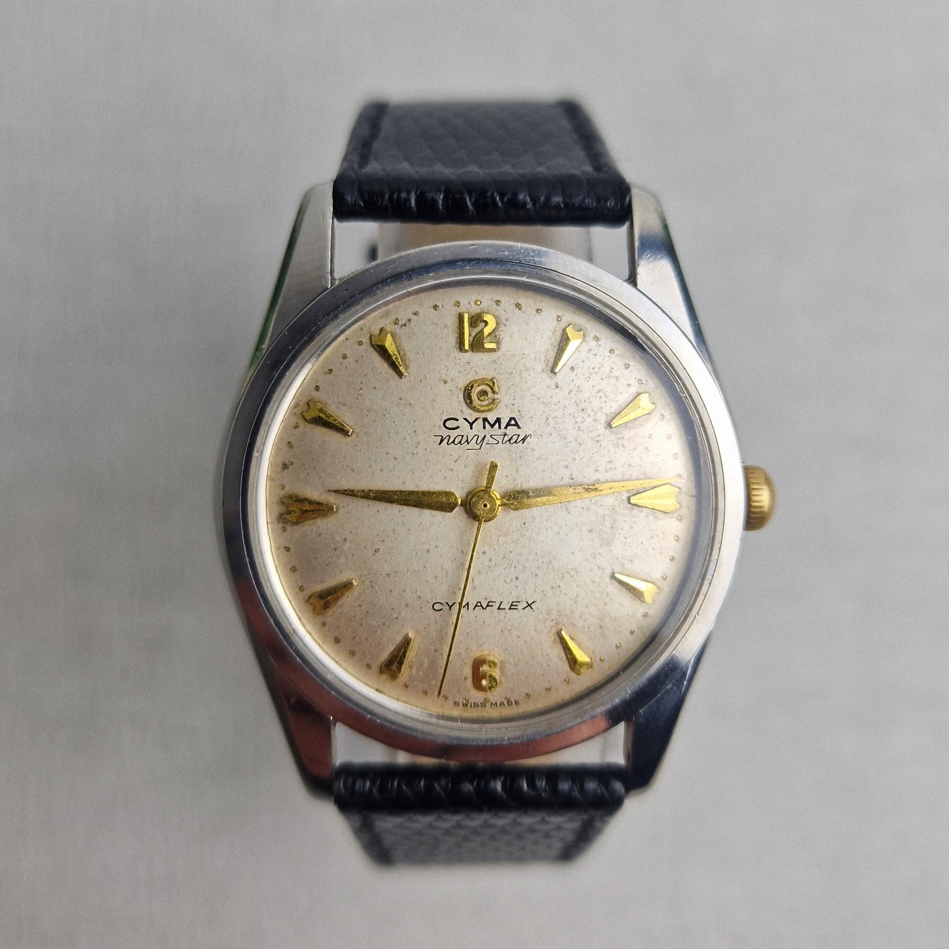 1950s Cyma Navystar Ref. 2-5678-6 featuring a 34mm stainless steel case, silvered dial with arrowhead markers, and black leather strap. Classic manual-wind Swiss watch.