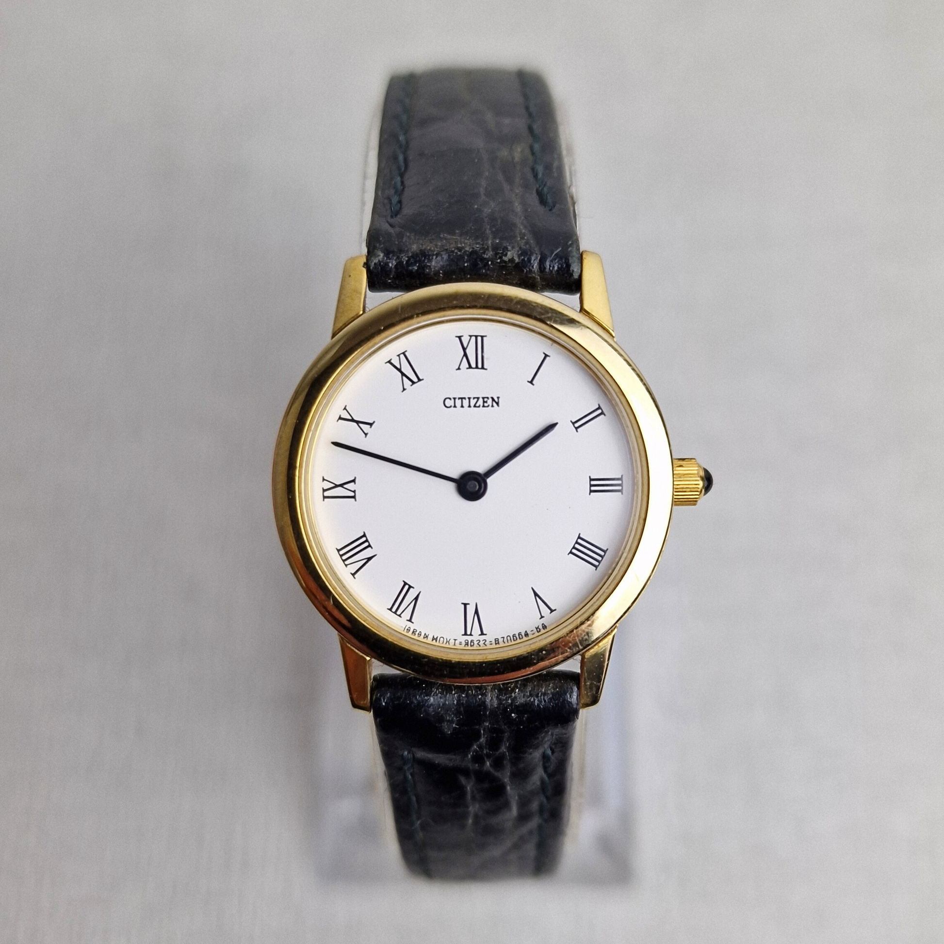 Front view of a vintage Citizen ladies watch with a gold-plated case, Roman numeral white dial, black leather strap, and cabochon crown.